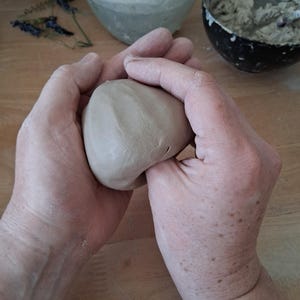 Könnte beinhalten: Hände formen einen Ball aus hellbraunem Ton. Der Ton wird zwischen den Handflächen geformt, mit anderen Töpferwerkzeugen und -materialien im Hintergrund. Die Szene deutet auf eine Töpferei oder einen Bildhauerworkshop hin.