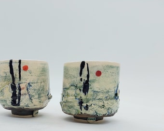 May include: Two handmade ceramic cups with a textured, off-white surface. Each cup features black brushstrokes, a red dot, and blue accents. The cups have a unique, organic shape and sit on a dark base.