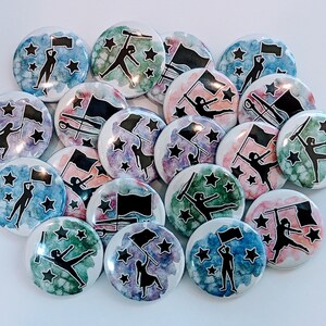 May include: A collection of 17 watercolor painted pinback buttons featuring silhouettes of color guard members with flags and stars. The buttons are in various shades of blue, green, pink, and purple.