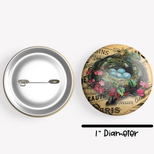 May include: A 1-inch diameter button featuring a vintage illustration of a bird's nest with blue eggs. The nest is surrounded by pink flowers and green leaves. The background is a faded, yellowed piece of paper with French text that reads "Beaut&eacute; de Paris" and "Nouvelles Galeries".