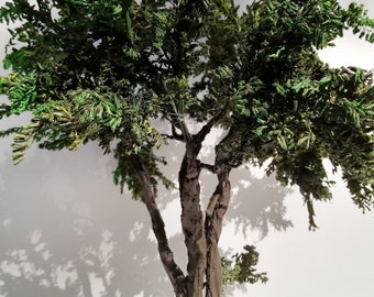 Könnte beinhalten: Ein kleiner künstlicher Baum mit braunem Stamm und grünen Blättern. Der Baum steht vor einer weißen Wand.