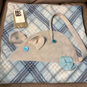 May include: A light blue and white plaid fleece blanket featuring a gray felt mouse applique with a bright blue button eye, a small blue bell attached to the tail, and a separate clear plastic bag containing dried catnip with a white label.