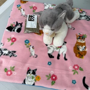 May include: A pink fleece blanket featuring a repeating pattern of various cats and colorful flowers, accompanied by a plush grey and white cat toy and a small clear plastic bag containing dried catnip with a white label attached to the top.