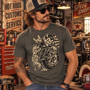 Pode incluir: Uma t-shirt cinzenta escura de manga curta com um gráfico de uma personagem de diabo de desenho animado montada num pneu. O design inclui o texto Hot Rods, Fuel Passion Freedom e Speed Shop, juntamente com uma bandeira axadrezada e um logótipo Mayobooks Garage.