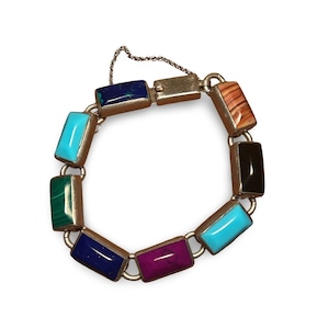 May include: A silver-toned bracelet featuring rectangular gemstone links in various colors, including turquoise, malachite, and a deep purple. The bracelet has a chain closure and is displayed against a white background.