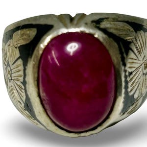 May include: A silver ring with a large, oval, deep red stone. The ring's band features etched floral designs and dark accents. The stone is the focal point, showcasing a polished, vibrant color. The ring is a statement piece.