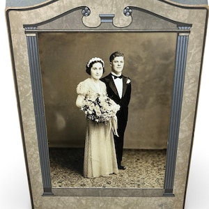 May include: Vintage framed photograph of a couple in formal attire. The black and white image shows a woman in a wedding dress holding a bouquet and a man in a tuxedo. The frame is a light color with decorative elements.