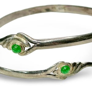 Puede incluir: Brazalete de puño plateado con dos elementos decorativos, cada uno con una gema verde brillante. El brazalete tiene un diseño abierto. Las gemas son redondas y están engastadas en engastes de plata ornamentados.