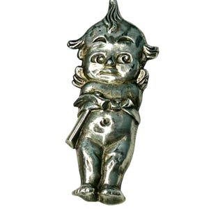 May include: A vintage silver-toned figurine of a cherubic figure with wings, a bow, and a stylized hairstyle. The figurine has a smiling face and is standing upright. The metal has a slightly tarnished appearance, suggesting age.