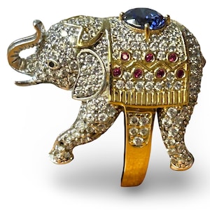 Peut inclure: Bague en forme d'éléphant doré, ornée de cristaux transparents. La trompe et les oreilles sont argentées. Un grand saphir bleu orne le dos, avec des pierres rouges et des détails dorés. La monture est dorée.