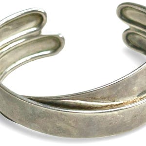 May include: A silver-toned cuff bracelet featuring a wide, smooth band that overlaps at the centre. The design includes an open back with two rounded, parallel ends that curve inward, creating a continuous, fluid shape with a polished metallic finish.