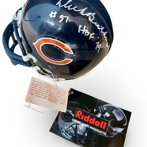 Puede incluir: Un casco de fútbol americano azul marino de los Chicago Bears con el logotipo del equipo y la firma de un jugador. El casco tiene una etiqueta blanca con texto y una etiqueta negra con el logotipo de Riddell.