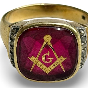 May include: A gold ring with a large, square, faceted, deep red stone. The stone features a gold Masonic symbol. The ring has a gold band and is accented with small, dark stones on each side of the red stone.