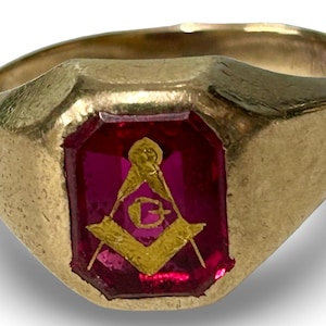 May include: A gold-colored ring with a rectangular, ruby-red stone. The stone features a gold-colored Masonic symbol. The ring has a rounded band and a slightly textured surface.