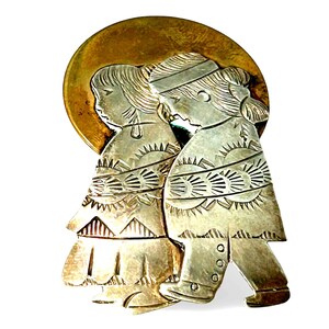 May include: A silver-toned metal brooch featuring two figures in traditional clothing, set against a gold-colored circular backdrop. The figures are intricately detailed with etched patterns, suggesting a cultural or historical theme. The brooch is likely a decorative accessory.