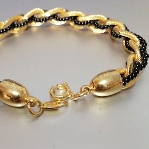 May include: A gold and black braided bracelet with a clasp closure. The bracelet is made of small beads and has a shiny finish.