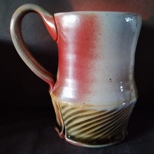May include: A handcrafted ceramic mug with a distinctive appearance. The mug showcases a red and white gradient on the upper section, blending into a textured, brown and tan pattern at the base. The handle matches the colour scheme, and the mug has a glossy finish.