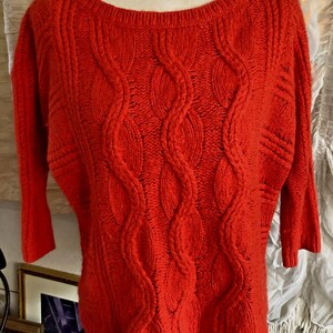 May include: Bright red sweater with a round neckline and three-quarter sleeves. The sweater features a cable knit design down the center and on the sleeves. The sweater is displayed on a mannequin.