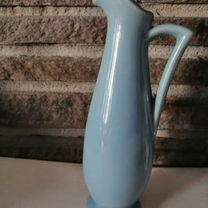 May include: A light blue ceramic pitcher with a slender body and a curved handle. The pitcher has a glossy finish.