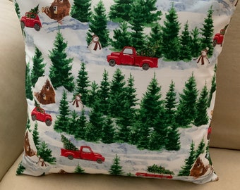 May include: A white throw pillow with a red truck and Christmas tree pattern. The truck is driving through a snowy forest with small houses and snowmen.