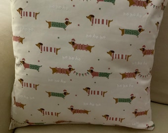 May include: White throw pillow with a repeating pattern of brown dachshunds wearing red and green sweaters. The dachshunds are wearing Santa hats and the text "ho ho ho" is repeated throughout the pattern.