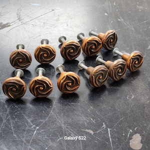 May include: A collection of ten bronze-colored cabinet knobs with a spiral design. Each knob has a round face with a detailed pattern and a silver-colored stem. The knobs are arranged on a dark, textured surface.