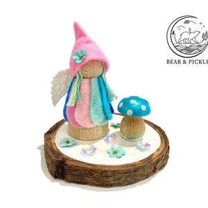 May include: A fairy ornament with a pink hat and colourful hair, wings, and clothing. A blue mushroom with white spots sits beside the fairy on a wooden base decorated with flowers. The Bear & Pickle logo is in the upper right corner.