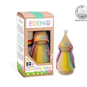 May include: A wooden peg doll named Eden, wearing a tan pointed hat with colourful flower accents and a multi-coloured felt cape with purple yarn hair. The doll is displayed next to its rectangular cardboard packaging featuring the text The Cornish Creative Piskie.