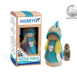 May include: A wooden peg doll figurine named Merryn, the Cornish Water Piskie, wearing a teal felt hood with a gold glitter crown, yellow yarn hair, and a heart-shaped sequin. It comes in a cardboard box with a small glass bottle of sea glass.
