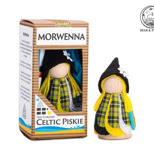 May include: A small wooden doll with yellow hair, a black hat with a white flower, and a yellow, black and red tartan scarf. The doll is packaged in a cardboard box with the words "The Cornish Celtic Piski" and "Morwenna".