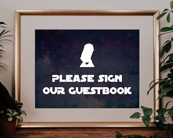 May include: Framed print with a gold border featuring a white silhouette on a dark blue and purple galaxy background. The text "PLEASE SIGN OUR GUESTBOOK" is displayed in white. The print is on a wooden surface with plants.