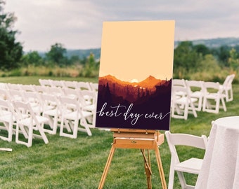 May include: A sign on a wooden easel features a mountain landscape in orange and purple, with the words "best day ever" in white script. The sign is displayed outdoors on grass, with rows of white chairs in the background, suggesting a wedding.