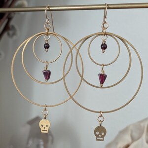 Puede incluir: Pendientes de aro dorados con tres círculos anidados. Cada pendiente presenta un pequeño dije de calavera, una cuenta de piedra preciosa de color rojo oscuro y una piedra preciosa en forma de lágrima. Los pendientes se exhiben en una barra dorada.