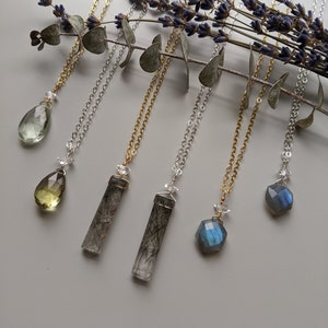 May include: Six necklaces with different crystal pendants, including a green teardrop, a yellow teardrop, a clear crystal with black inclusions, a blue crystal, and two grey crystals with black inclusions. The necklaces are on a white background with lavender and eucalyptus branches.