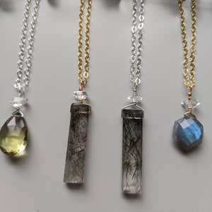 May include: Four necklaces with different crystal pendants. The pendants are a light green teardrop, a clear crystal with black inclusions, a clear crystal with black inclusions, and a blue labradorite crystal.