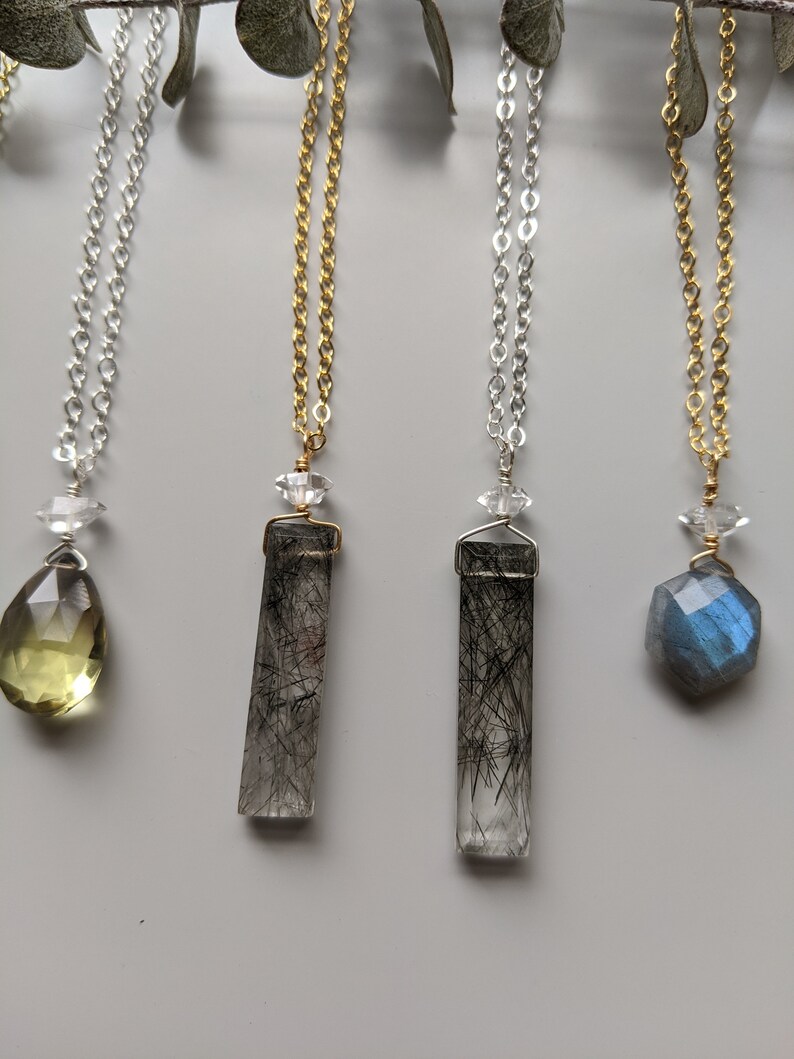 May include: Four necklaces with different crystal pendants. The pendants are a light green teardrop, a clear crystal with black inclusions, a clear crystal with black inclusions, and a blue labradorite crystal.