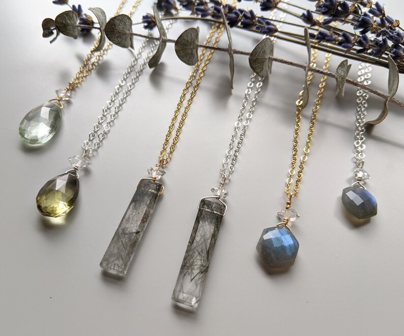 May include: Six necklaces with different crystal pendants. The pendants include a teardrop-shaped green crystal, a teardrop-shaped yellow crystal, a long rectangular clear crystal with black inclusions, a long rectangular clear crystal with black inclusions, a hexagonal blue crystal, and a hexagonal blue crystal.