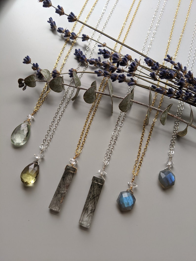 May include: Six necklaces with different crystal pendants, including a green teardrop, a yellow teardrop, a clear crystal with black inclusions, a blue crystal, and two grey crystals with black inclusions. The necklaces are on a white background with lavender and eucalyptus branches.