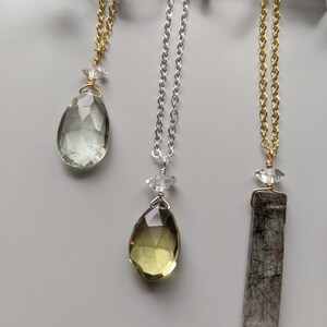 May include: Three necklaces featuring different coloured gemstones. The first necklace has a light green teardrop-shaped gemstone, the second necklace has a light yellow teardrop-shaped gemstone, and the third necklace has a clear rectangular gemstone with black inclusions.