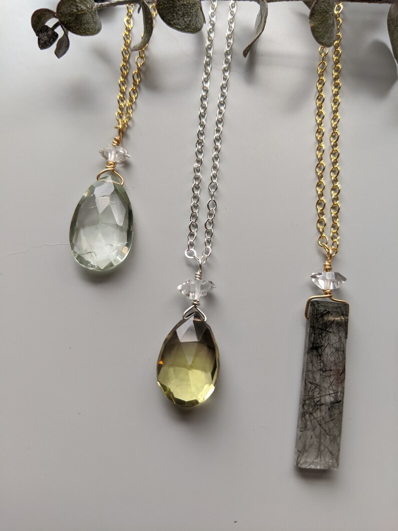 May include: Three necklaces featuring different coloured gemstones. The first necklace has a light green teardrop-shaped gemstone, the second necklace has a light yellow teardrop-shaped gemstone, and the third necklace has a clear rectangular gemstone with black inclusions.
