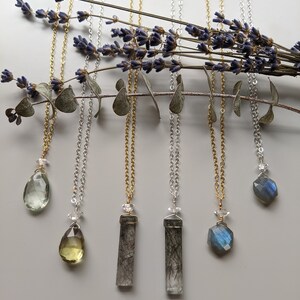 May include: Five necklaces with different gemstone pendants. The pendants include a green teardrop, a yellow teardrop, two clear rectangular stones with black inclusions, and a blue faceted stone. The necklaces are on a white background with lavender and eucalyptus branches.