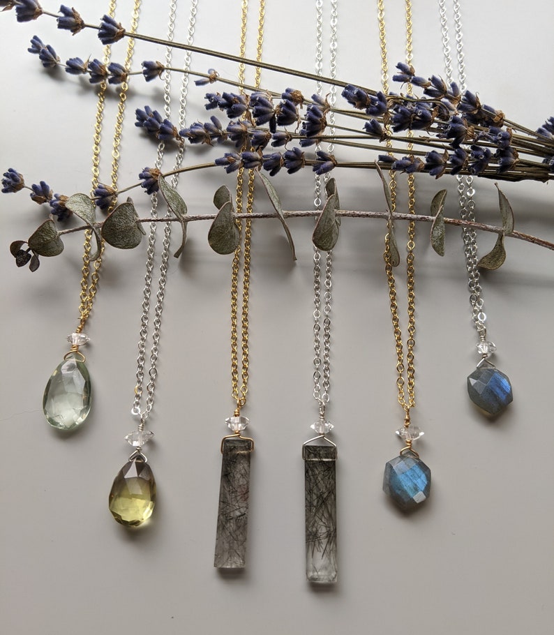 May include: Five necklaces with different gemstone pendants. The pendants include a green teardrop, a yellow teardrop, two clear rectangular stones with black inclusions, and a blue faceted stone. The necklaces are on a white background with lavender and eucalyptus branches.