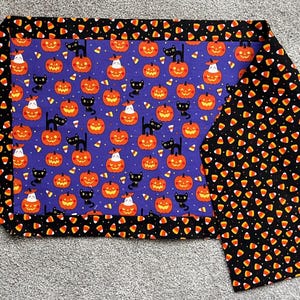 May include: A rectangular table runner featuring a central purple panel with a repeating pattern of orange jack-o'-lanterns, black cats, and candy corn. The border is black fabric with a dense, repeating print of orange, yellow, and white candy corn.