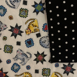 May include: A white fabric with a repeating pattern of cartoon cats in various poses, along with starburst and square designs. A black fabric with white polka dots is on the right side.
