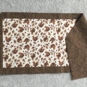 May include: A brown table runner with a white background featuring a repeating pattern of squirrels and leaves. The runner has a brown border.