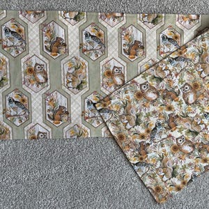 May include: A rectangular fabric table runner featuring a repeating pattern of woodland animals including owls, squirrels, and birds within hexagonal frames on a light green background, with a folded corner showing a dense floral and animal print.
