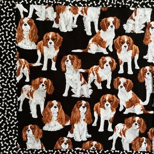 May include: A black fabric with a repeating pattern of Cavalier King Charles Spaniel dogs in shades of brown, white, and black. The border features a white bone pattern on a black background. The dogs are in various poses.