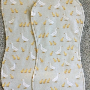 May include: Two baby burp cloths with a light blue background. The cloths are patterned with white geese and yellow ducklings. The edges are rounded and the cloths are made of soft fabric.