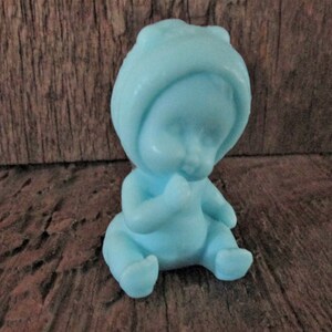 May include: A small blue baby figurine with a hooded hat, sitting with its hands clasped in front of its face.