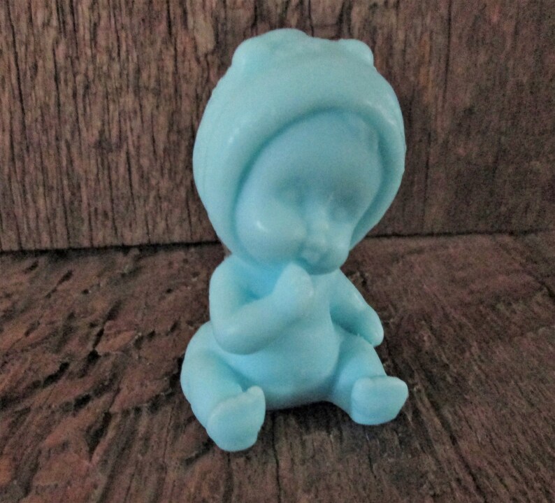 May include: A small blue baby figurine with a hooded hat, sitting with its hands clasped in front of its face.